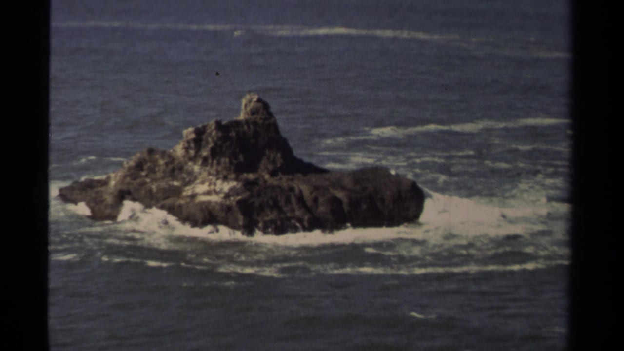 Dark rocky island in the ocean with foam and waves around it.