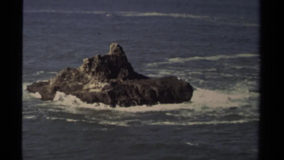 Distant Mendocino, California home-movie view of a dark rock island ringed by white Pacific surf.