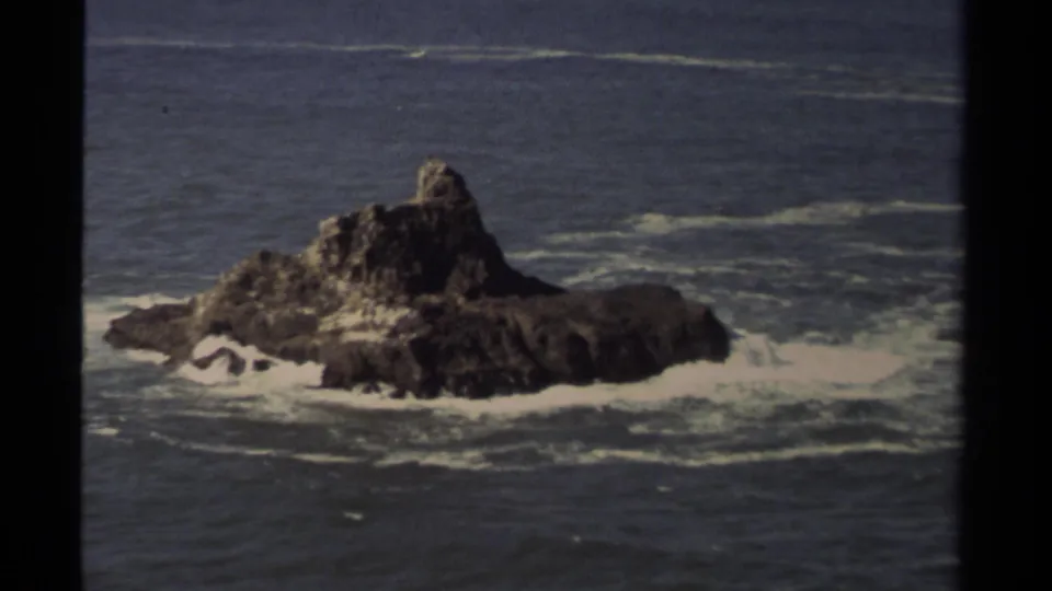 Vintage 8mm coastal shot showing a rugged Mendocino offshore outcrop with waves washing across its base.