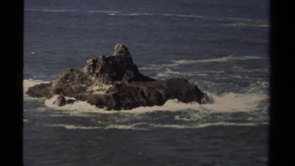 Mendocino 1980 home-movie frame of a jagged offshore rock set against blue-gray water and white surf.