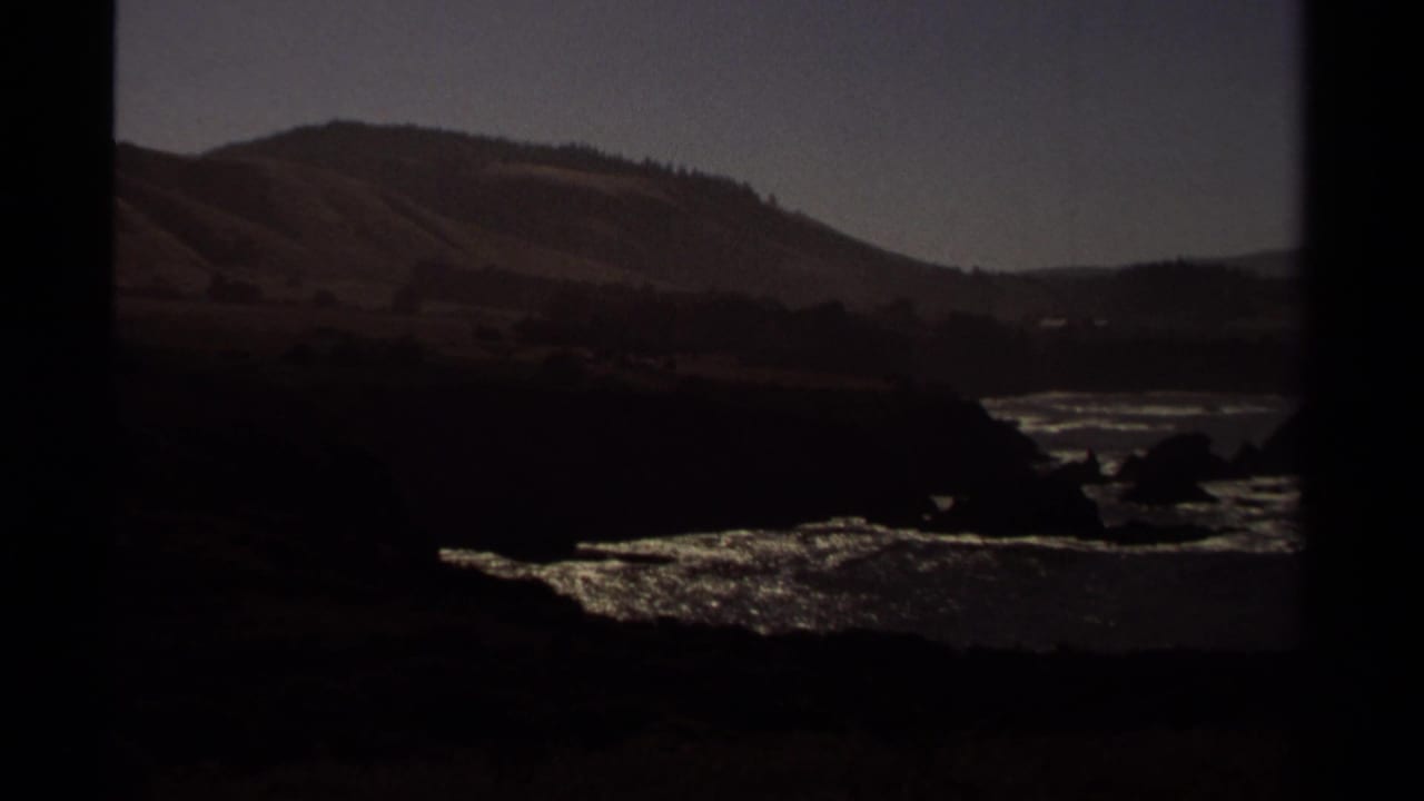 1980 home-movie frame of the Mendocino coast, with dark rocks, shimmering surf, distant hills, and visible film-frame edges.