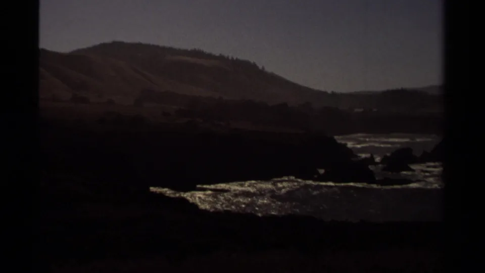 1980 home-movie frame of the Mendocino coast, with dark rocks, shimmering surf, distant hills, and visible film-frame edges.