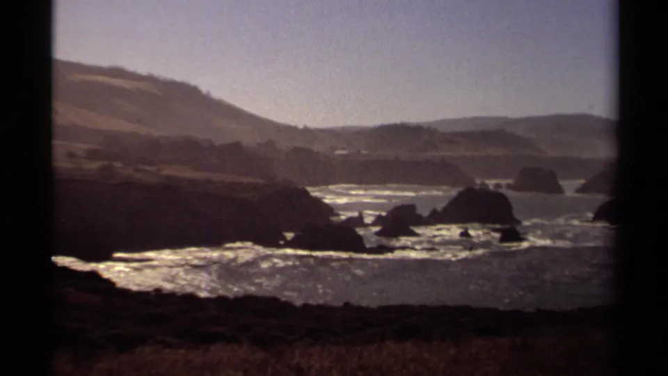 Vintage 8mm frame of the Mendocino shoreline, with offshore sea stacks, bright surf, hillside terrain, and dark gate edges.