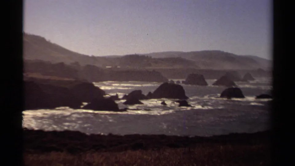Wide 1980 Mendocino coast frame with sea stacks in the water, low surf, grassy foreground, hazy hills, and soft Super 8 grain.