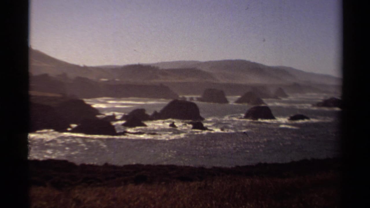 1980 8mm home-movie view of the Mendocino coast with offshore rock formations, breaking surf, hazy hills, and dry grass in the foreground.