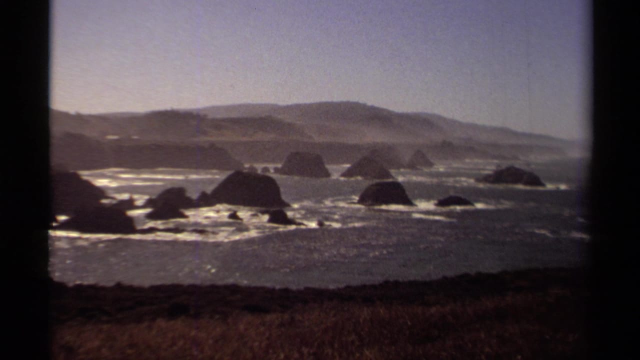 Offshore rocks and bright surf below rolling hills, seen from a grassy coastal overlook.