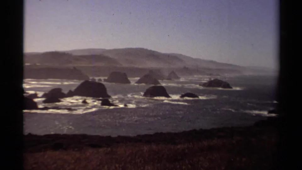 Grainy 8mm frame of the 1980 Mendocino shoreline, showing dark offshore rocks, white surf patterns, distant hills, and a dry grassy bluff.