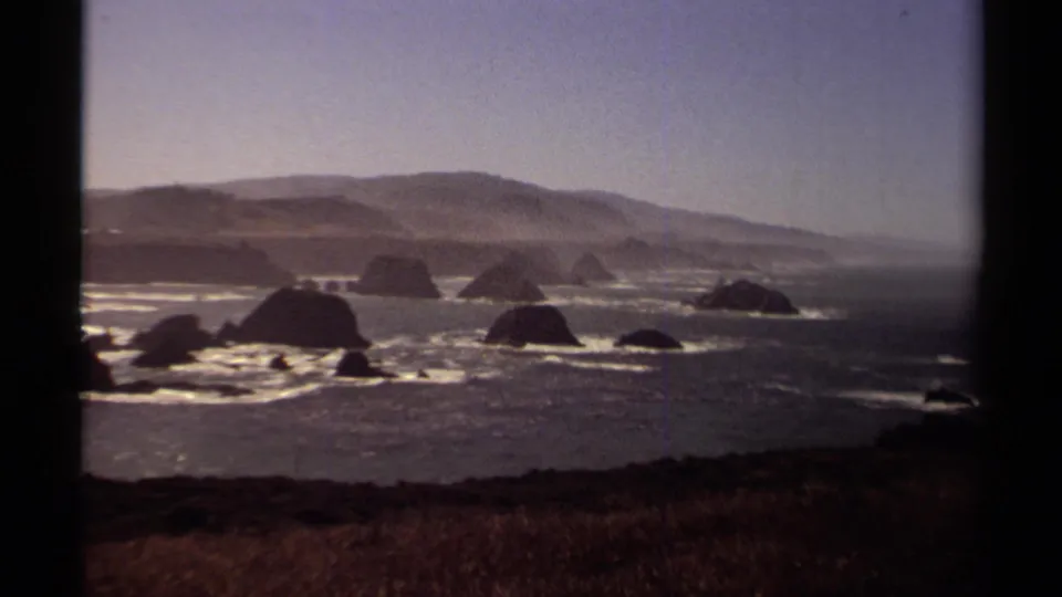 Final 1980 8mm view of the Mendocino coast, with offshore rock shapes, glinting water, low surf, rolling hills, and warm home-movie color.