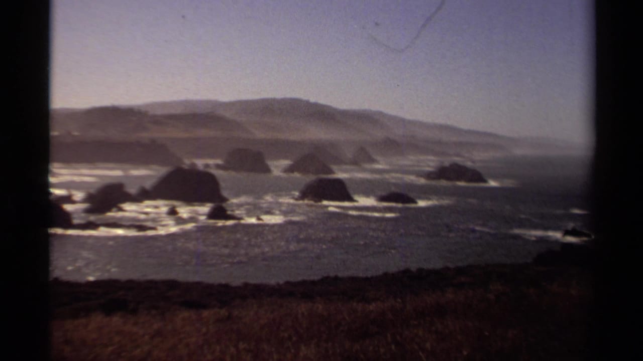 Rocky coastline with sea stacks, surf, distant hills, and dark film-frame borders.
