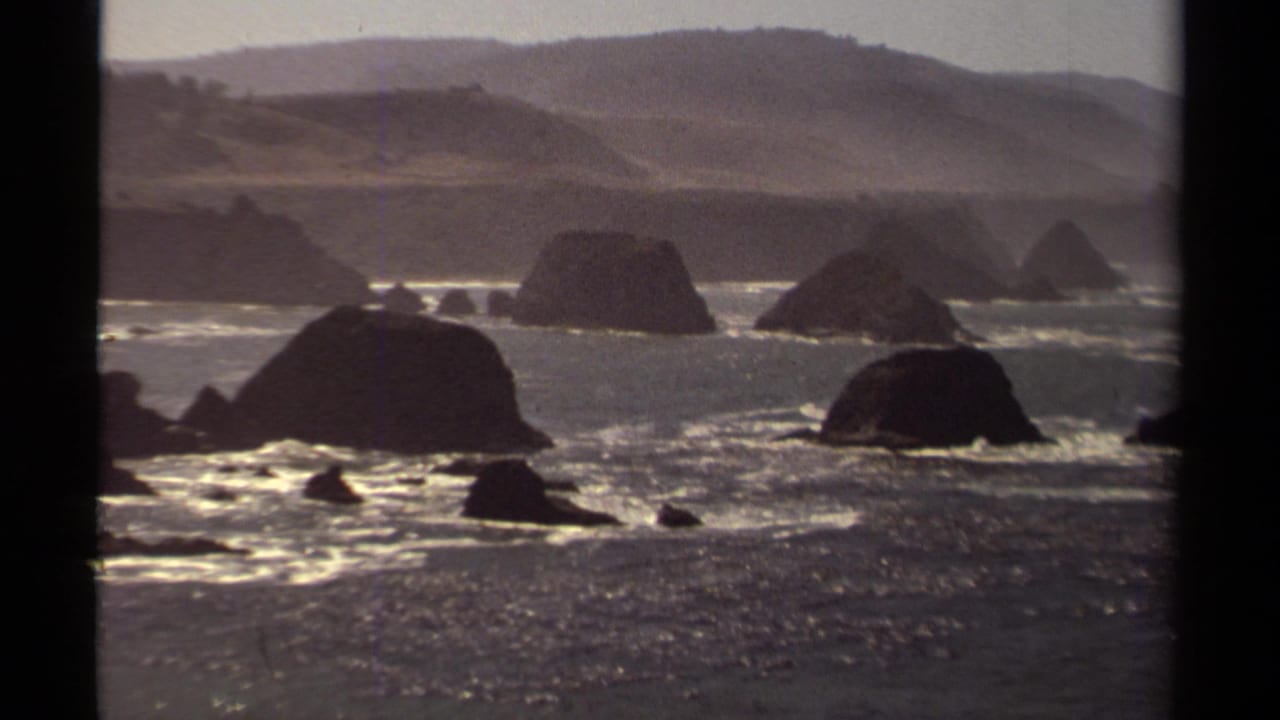 Mendocino, California coastal scene from 1980 8mm footage, showing large offshore rocks, bright breaking waves, and muted hills.