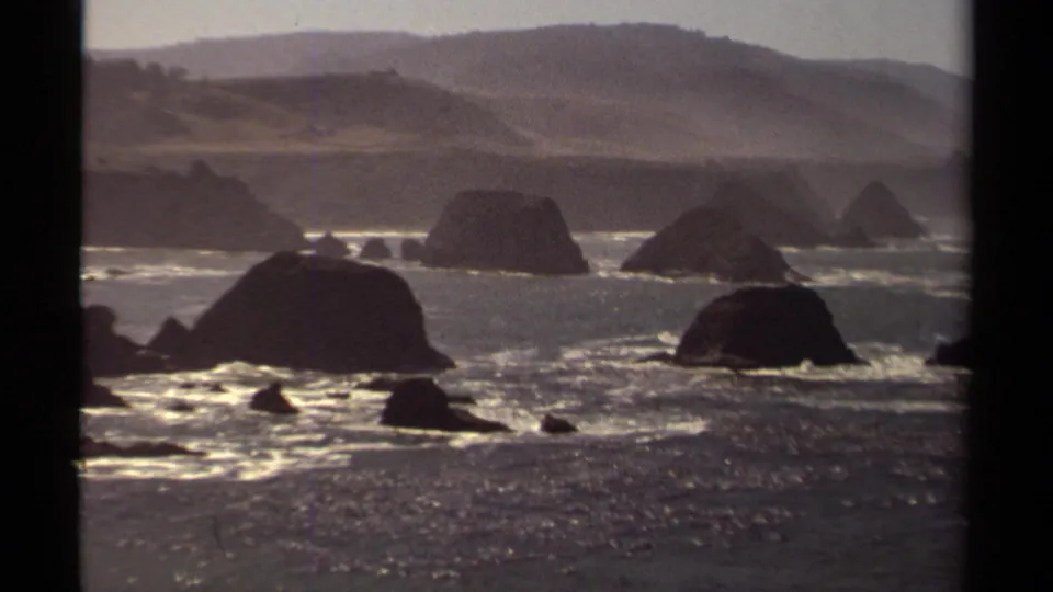 Mendocino, California coastal scene from 1980 8mm footage, showing large offshore rocks, bright breaking waves, and muted hills.