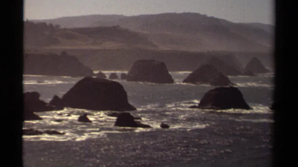Vintage 8mm frame of Mendocino sea stacks, churning ocean surface, bright surf bands, and distant hillside under soft haze.