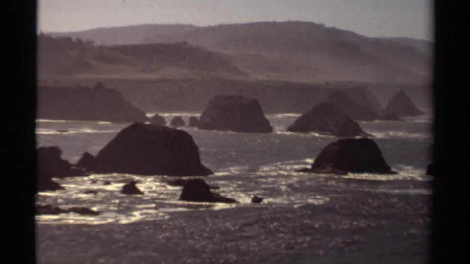 1980 home-movie view of the Mendocino shoreline with offshore rocks, layered surf, muted hills, and visible 8mm frame edges.