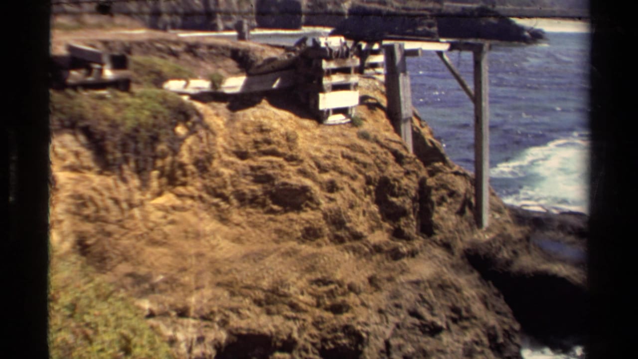 Rocky coastal cliff with surf below and a small wooden structure near the edge.