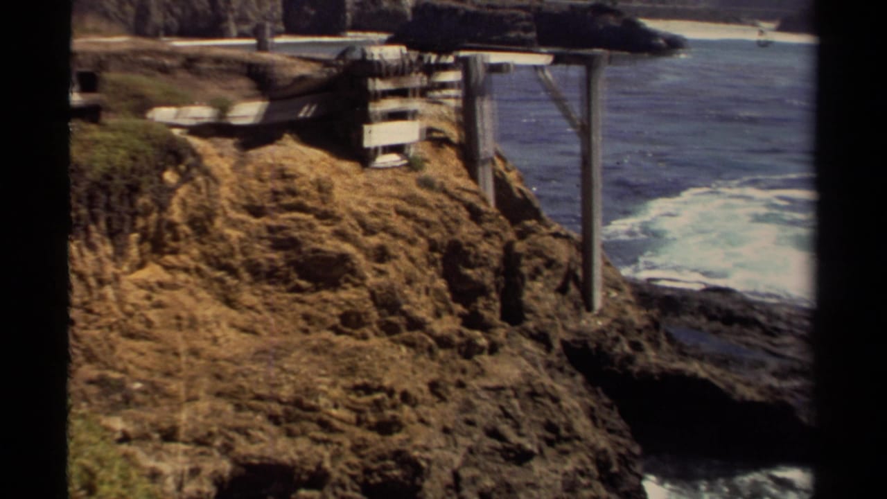 1980 Mendocino-area home-movie frame with cliffside wooden supports, dry coastal vegetation, dark rock, and waves breaking below.