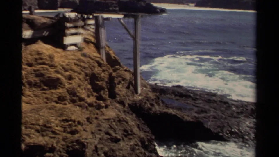 1980 Mendocino-area 8mm frame showing white surf, dark shoreline rocks, a cliff edge, and partial wooden supports at the waterline.