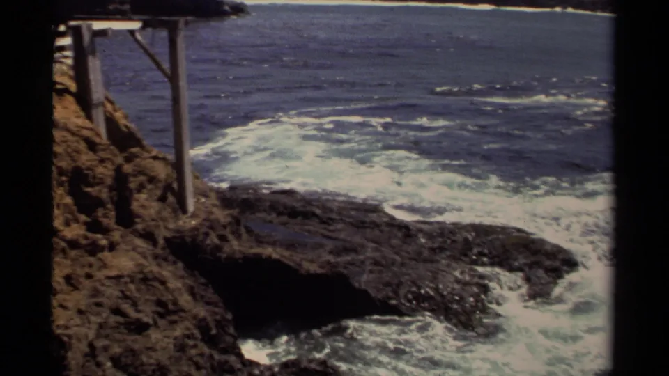 1980 Mendocino-area frame focusing on Pacific surf around dark rock ledges, with part of a wooden support structure at the cliff edge.