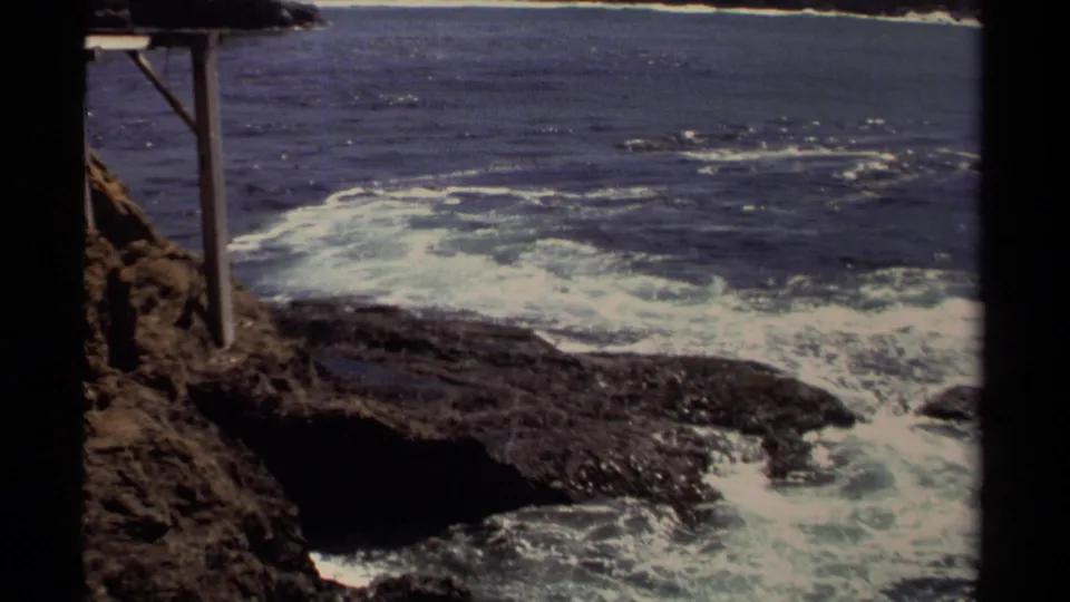 1980 8mm Mendocino coastal view with white surf washing over dark rocks near a wooden post structure.