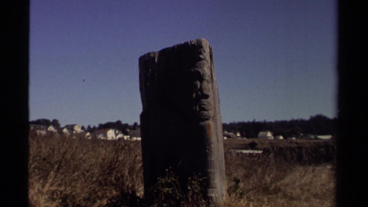 Weathered wooden post with a carved face stands in a dry Mendocino field, with white houses and blue sky in the background.