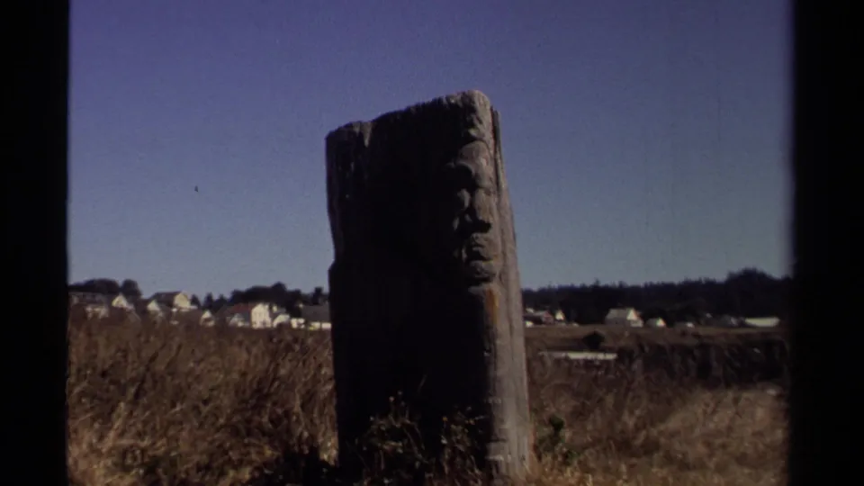 Weathered wooden post with a carved face stands in a dry Mendocino field, with white houses and blue sky in the background.