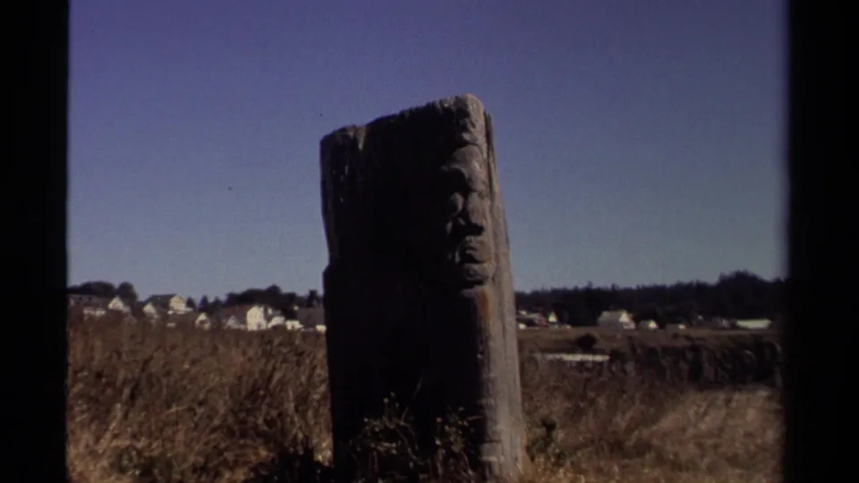 The carved wooden post appears closer in frame, set against dry Mendocino grassland and distant white houses in 1980 footage.