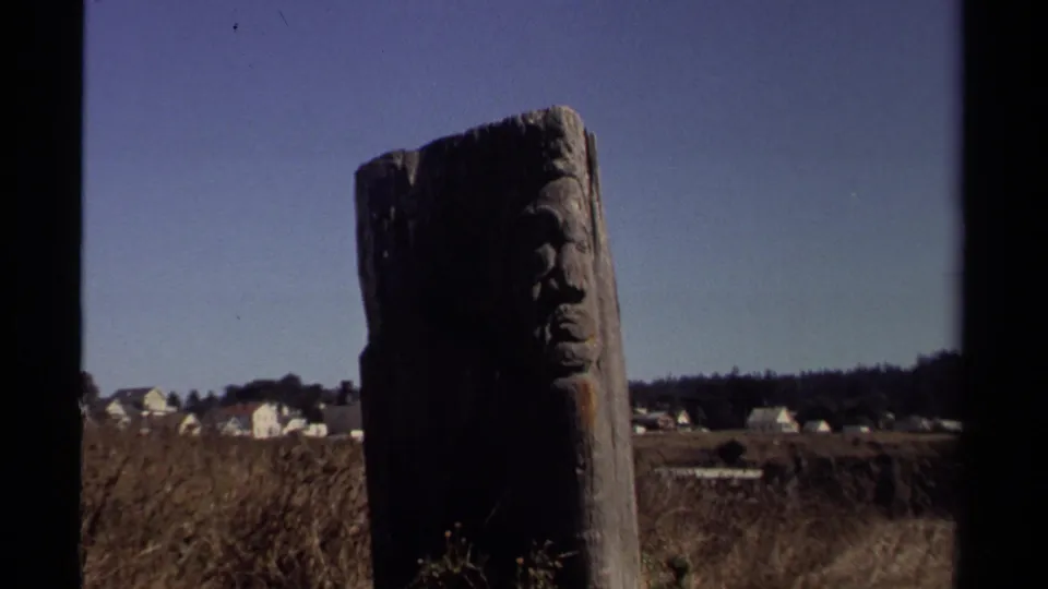 Weathered carved post remains centered in a dry Mendocino field, with small white houses and a wooded ridge beyond.