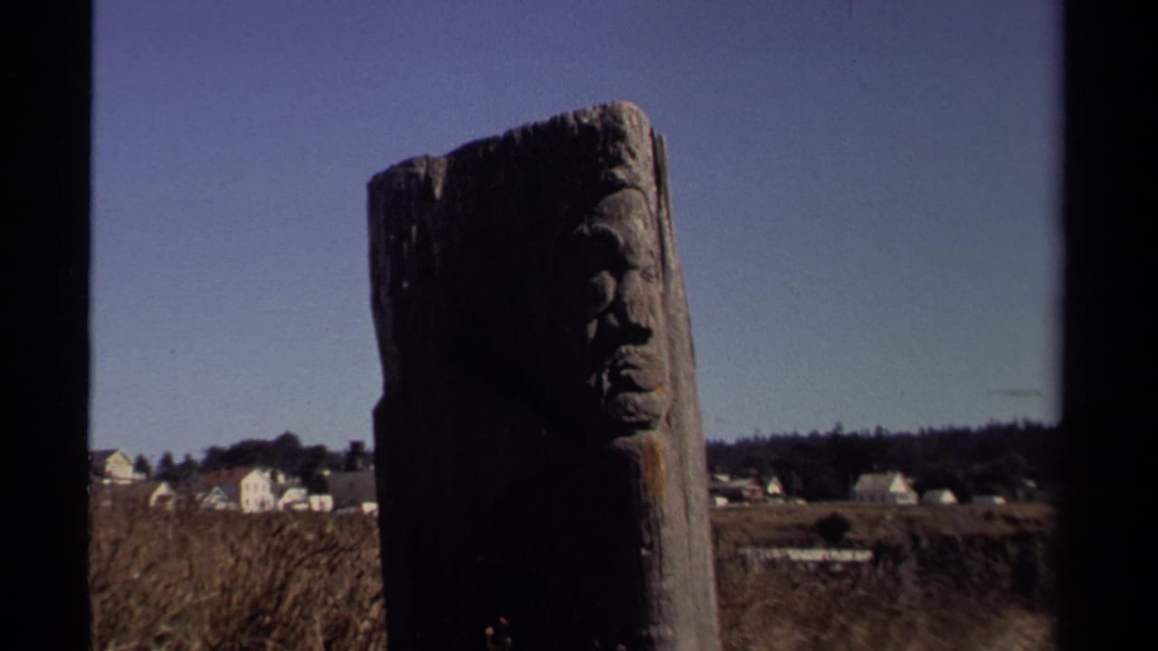 1980 Mendocino 8mm frame showing a weathered carved wooden pole in front of distant white houses and low vegetation.