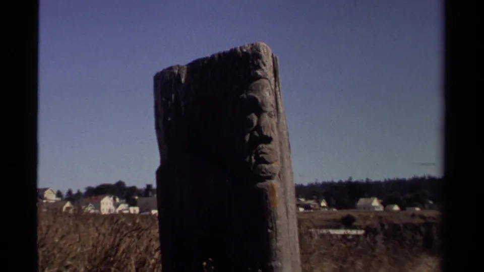 1980 Mendocino 8mm frame showing a weathered carved wooden pole in front of distant white houses and low vegetation.