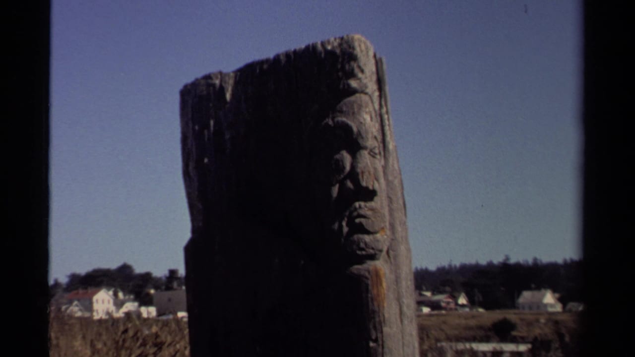 Carved wooden face on a tall weathered pole with houses and trees in the distance.