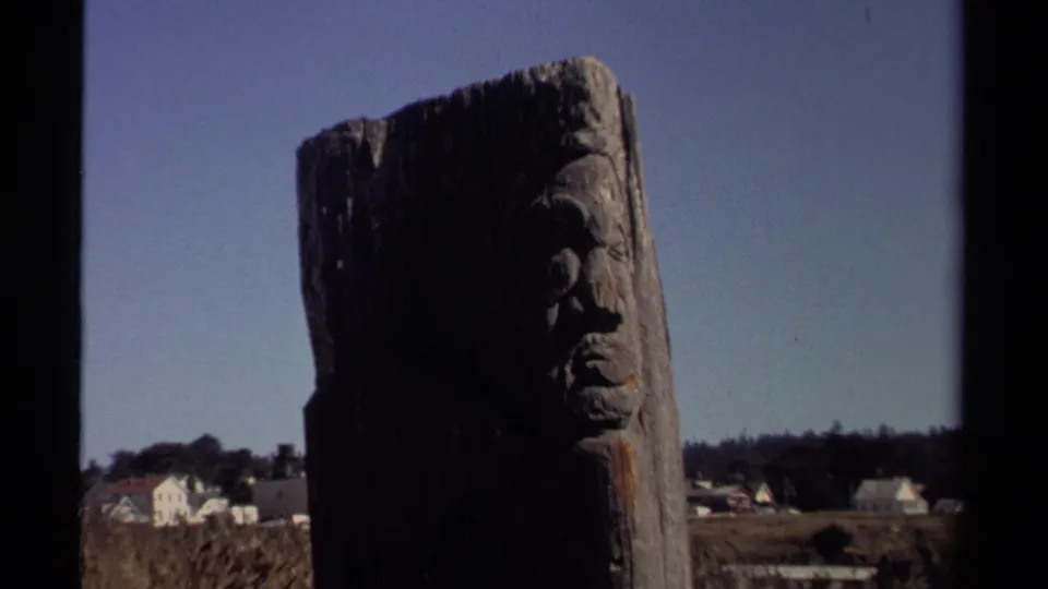 The Mendocino landmark view holds on the carved wooden pole, with a small town and dark tree line behind it.