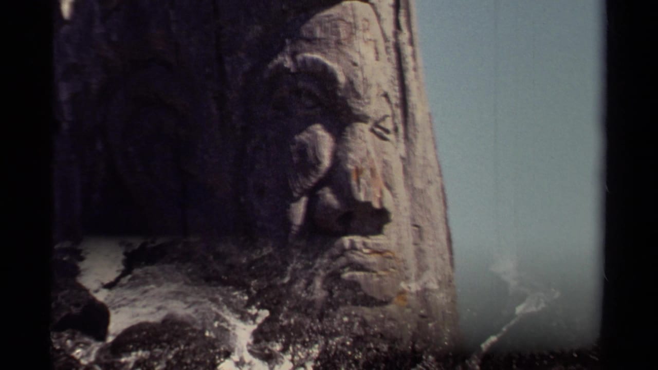 Carved wooden face above foaming surf and dark coastal rocks.