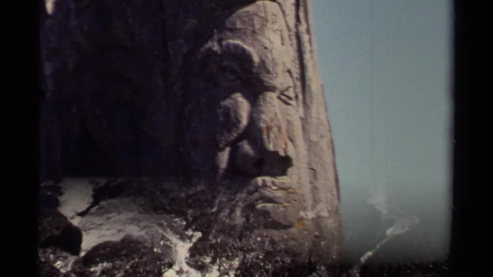 1980 8mm Mendocino-area frame with a weathered carved wooden face overlooking foaming waves on dark rocks below.