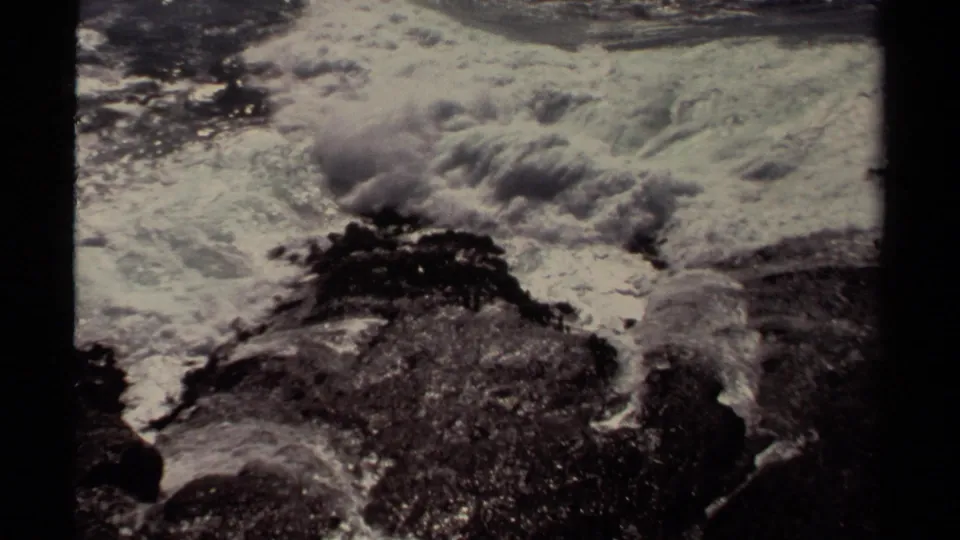 1980 8mm Mendocino-area frame showing white surf breaking across dark jagged rocks along the coastline.