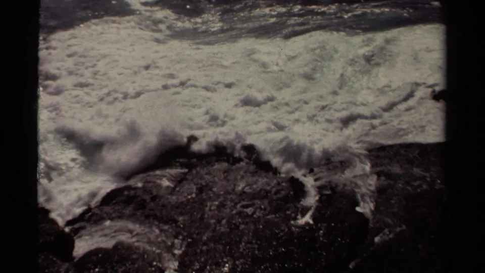 1980 8mm Mendocino-area frame with foaming ocean water rolling over dark shoreline rocks in close view.