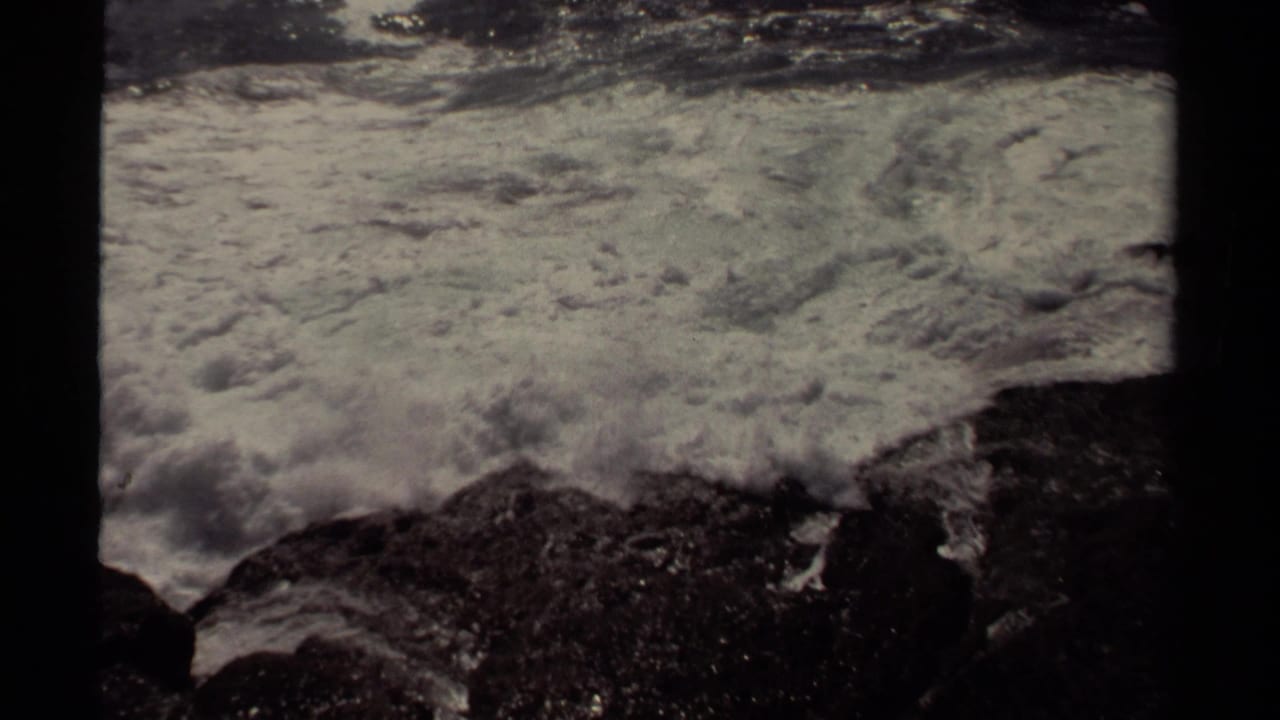 White ocean foam spreads over dark shoreline rocks within a black 8mm frame border.