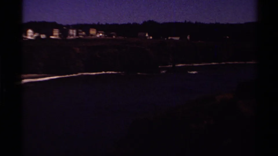 Color 1980 8mm view of a Mendocino coastal town across dark water, with wooded hills behind.