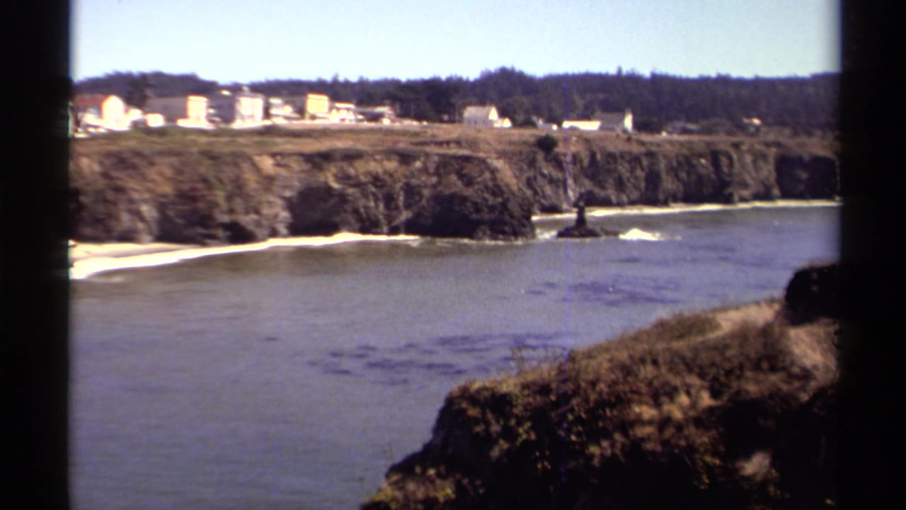 Rocky coastal cove with ocean water, small beach, cliffs, and distant houses.