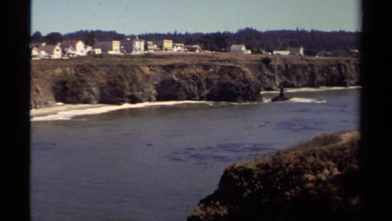 Mendocino coastal view with ocean, rugged bluff, narrow beach, distant houses, and dark trees in the background.