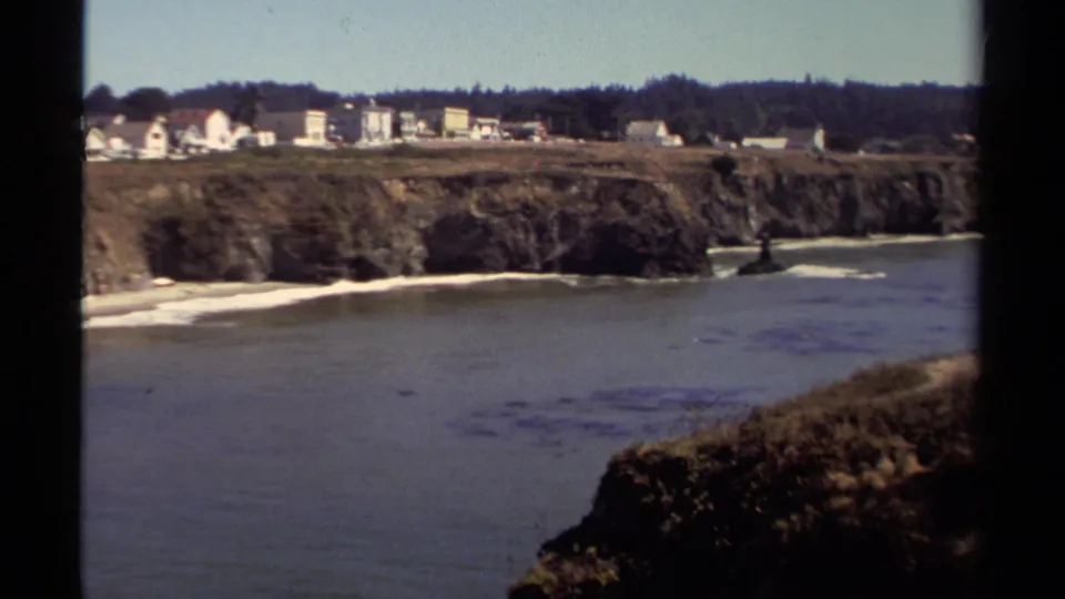 Mendocino coastal view with ocean, rugged bluff, narrow beach, distant houses, and dark trees in the background.
