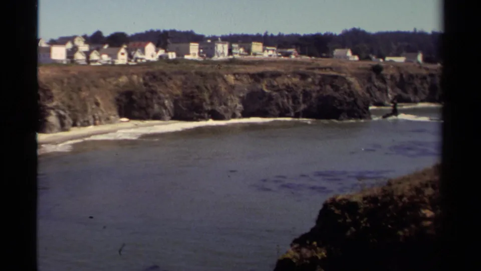 Vintage 8mm frame of the Mendocino shoreline, showing a beach cove, rock cliffs, ocean water, homes, and trees.