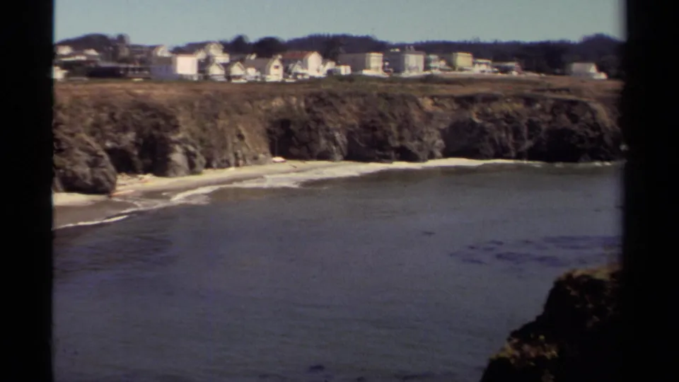 Mendocino 8mm coastal frame with a broad cove, rocky shoreline, pale beach, distant homes, and a wooded ridge.