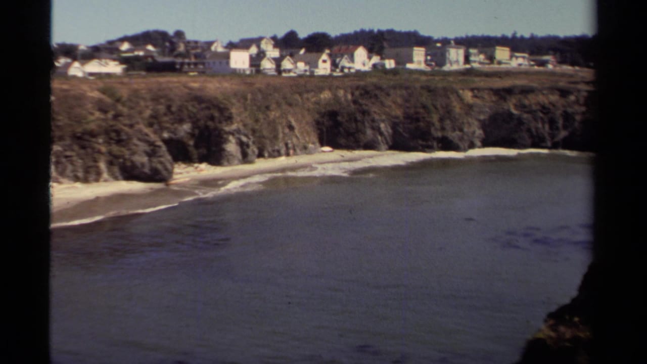 Elevated view of a sandy beach below cliffs with distant hillside houses.