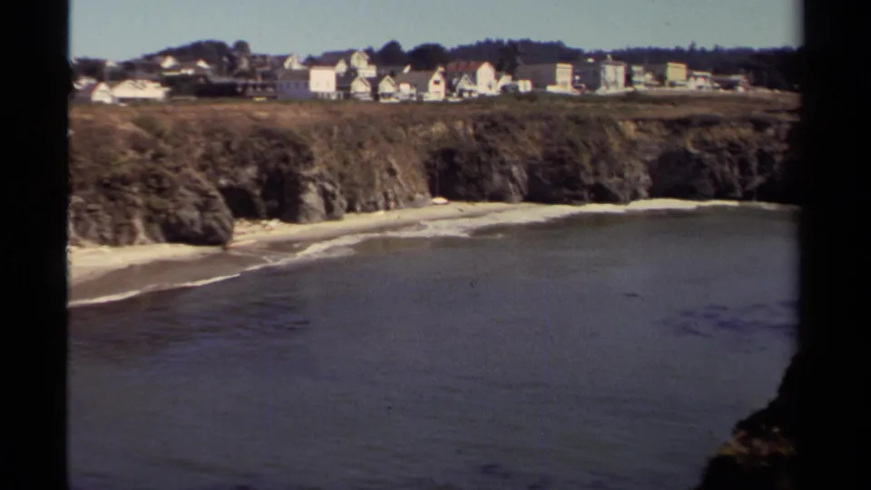 1980 Mendocino coastal view with calm water, a narrow beach, rugged cliffs, and houses along the bluff.