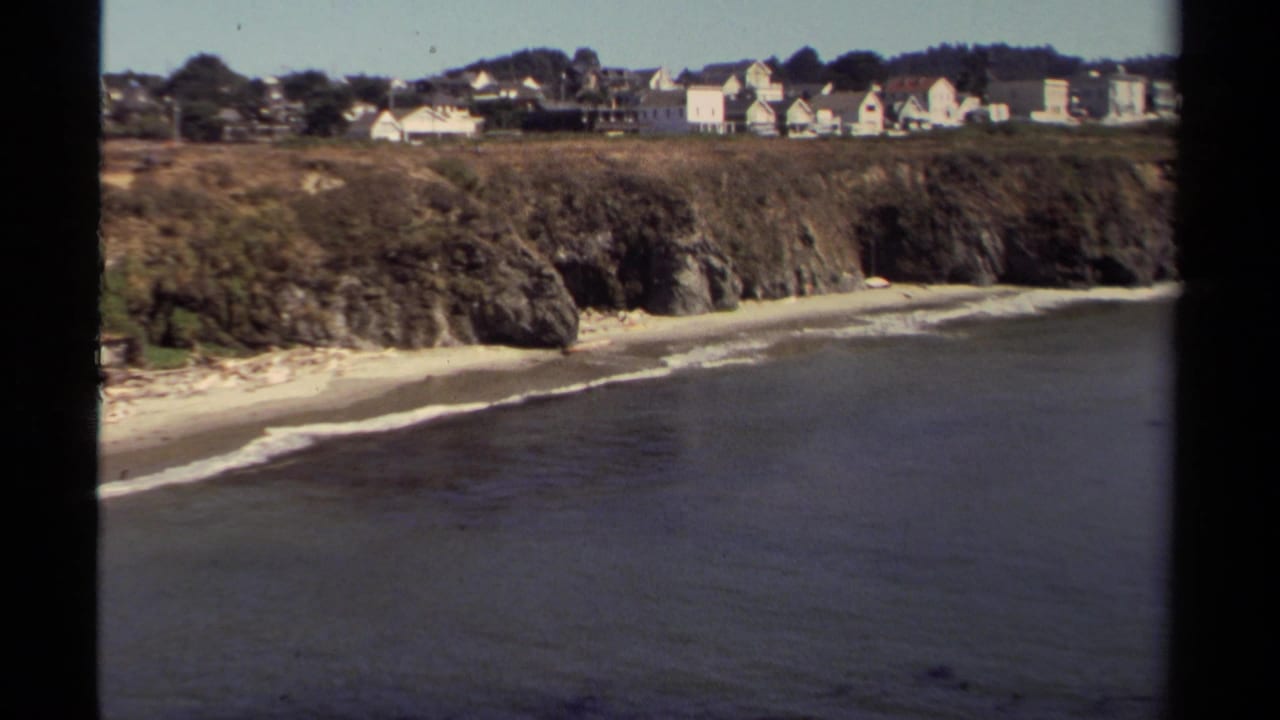 Elevated 8mm view of Mendocino shoreline, with gentle surf, cliffs, beach, and hillside homes in the distance.
