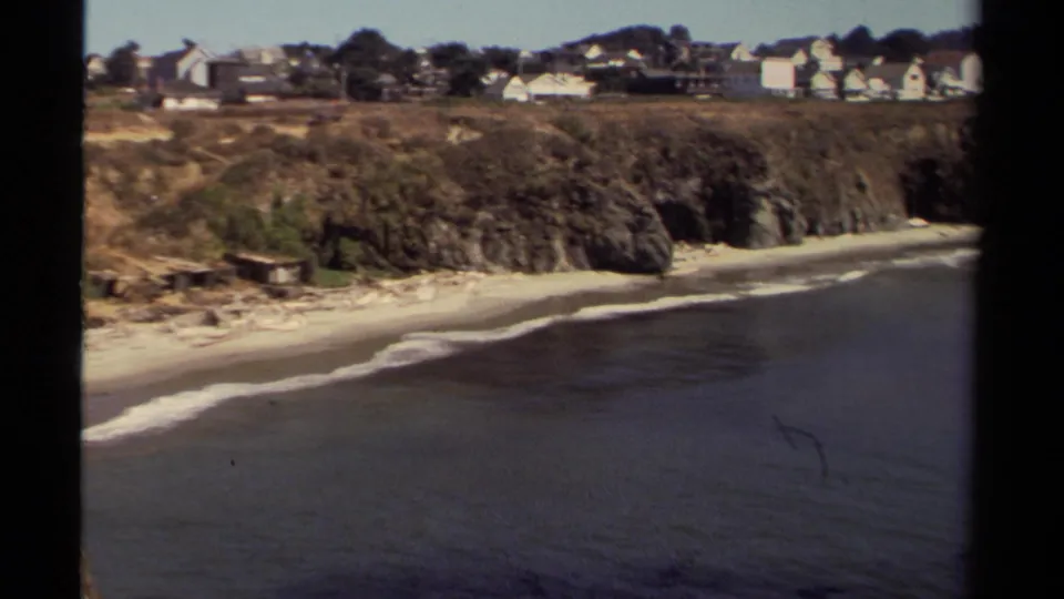 Mendocino beach cove seen from above, showing low surf, rocky bluffs, scrub vegetation, and distant homes.