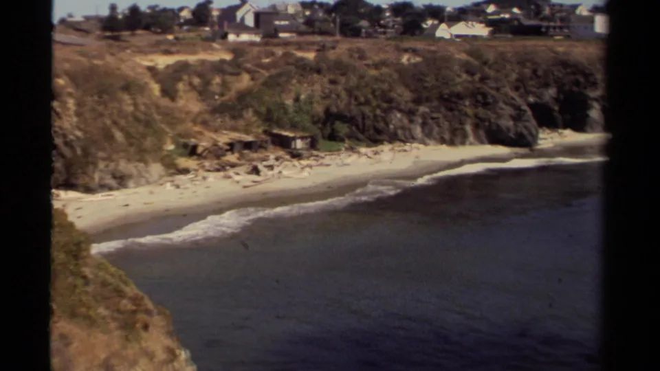Final elevated view of the Mendocino shoreline with gentle surf, sandy beach, rocky bluff, vegetation, and distant hillside buildings.