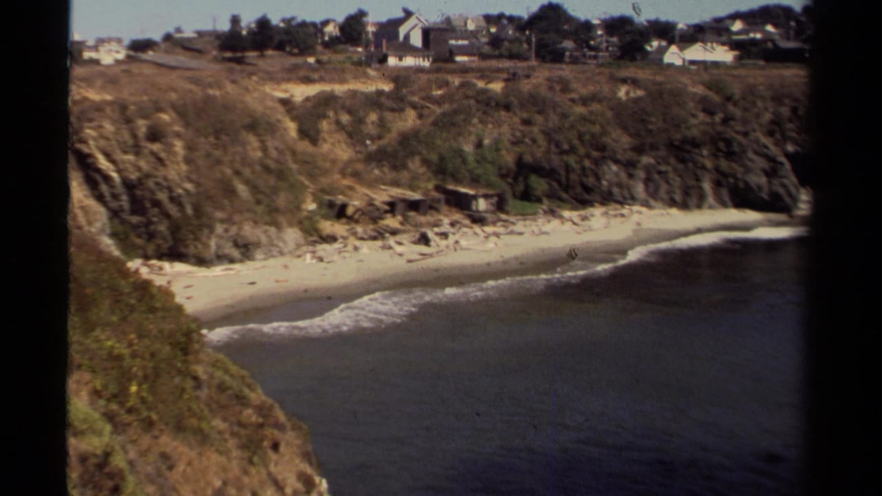 Mendocino coastal cove in 1980 8mm footage, showing surf, a sandy beach, bluff vegetation, and distant homes above the shoreline.