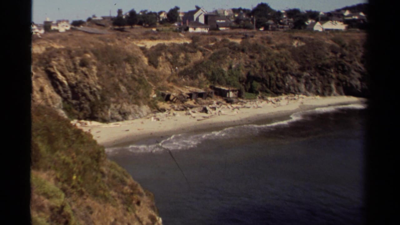 Coastal cove with surf breaking along the beach beneath grassy bluffs and houses.