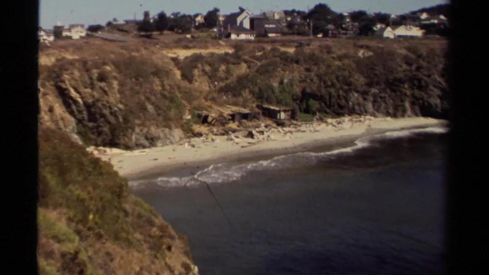 Vintage 8mm view of a Mendocino-area beach cove, with dark ocean water, white surf, eroded bluffs, and houses set back above.