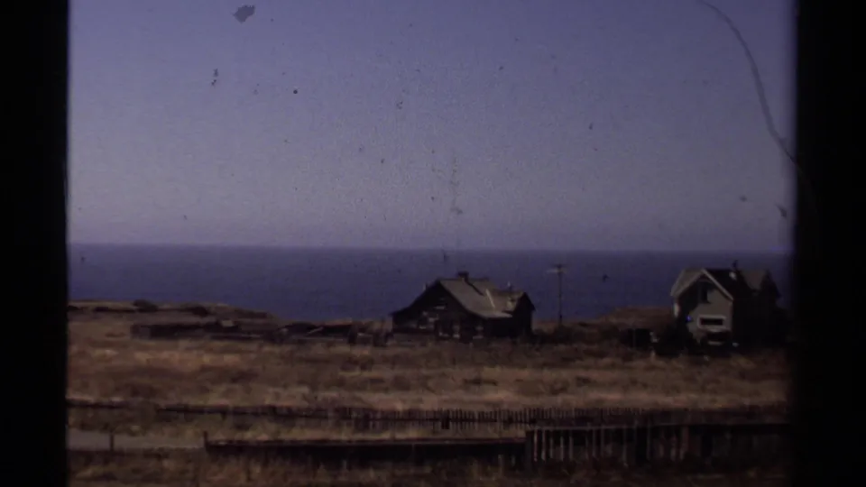 Mendocino coastal home-movie frame with houses, a fence, dry bluff grass, and the Pacific horizon under a hazy sky.